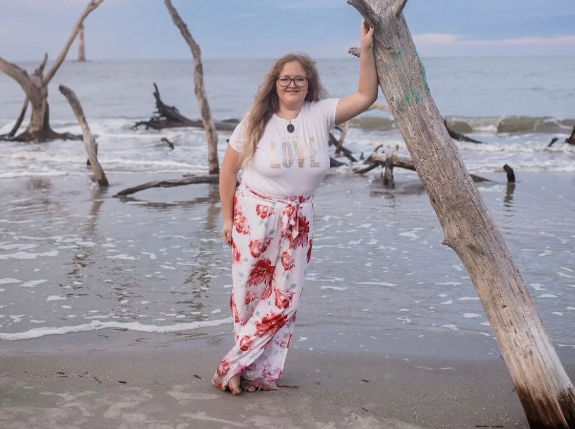 Mujer de pie en una playa sosteniendo un árbol de madera flotante, con olas del mar y árboles caídos al fondo.